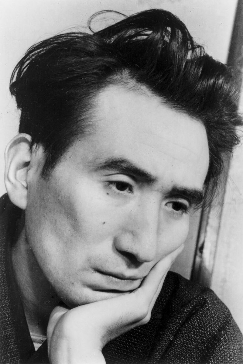 Close no rosto de um homem japonês. Ele tem cabelos longos, ao estilo dos anos 50. Tem uma mão apoiada no queixo e olha, de pálpebras baixas, para um ponto baixo. Foto em preto e branco.