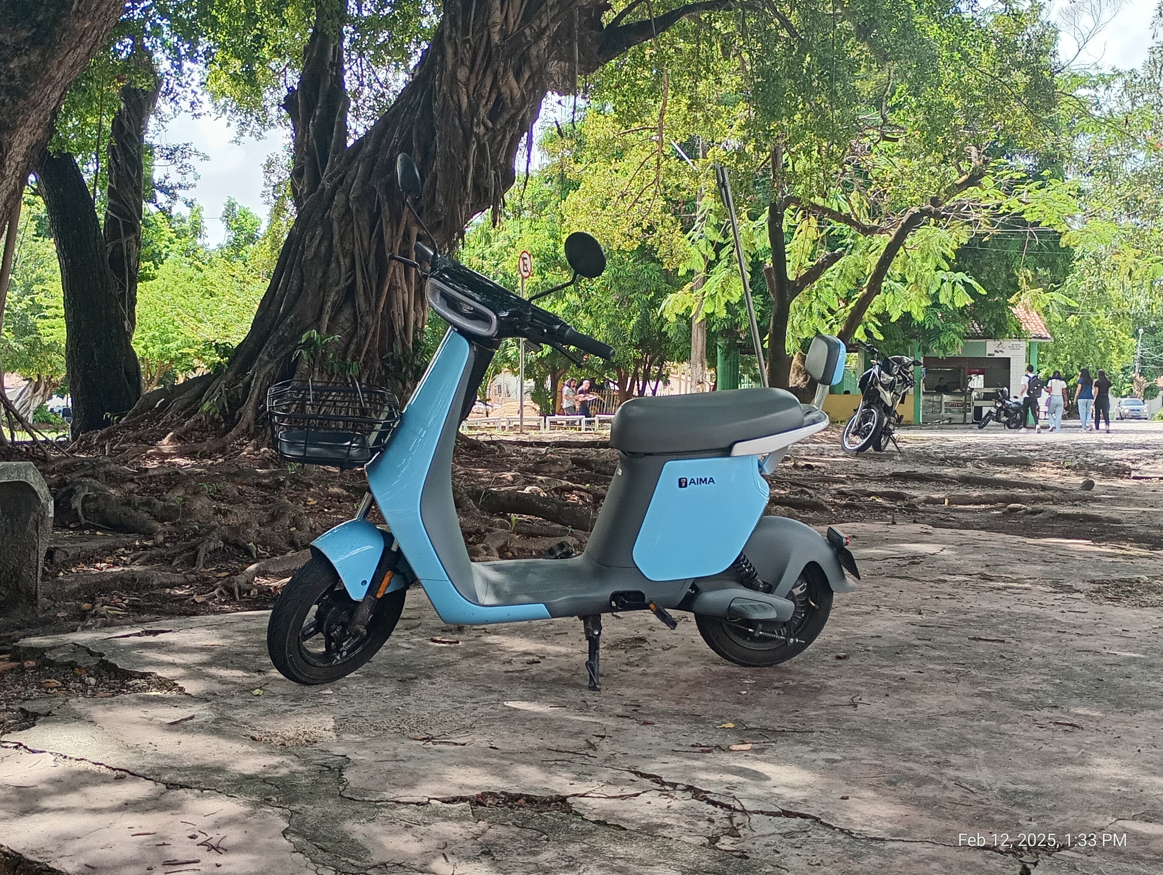 Foto de uma lambreta de cor azul-bebê. Ao fundo, um bosque universitário.