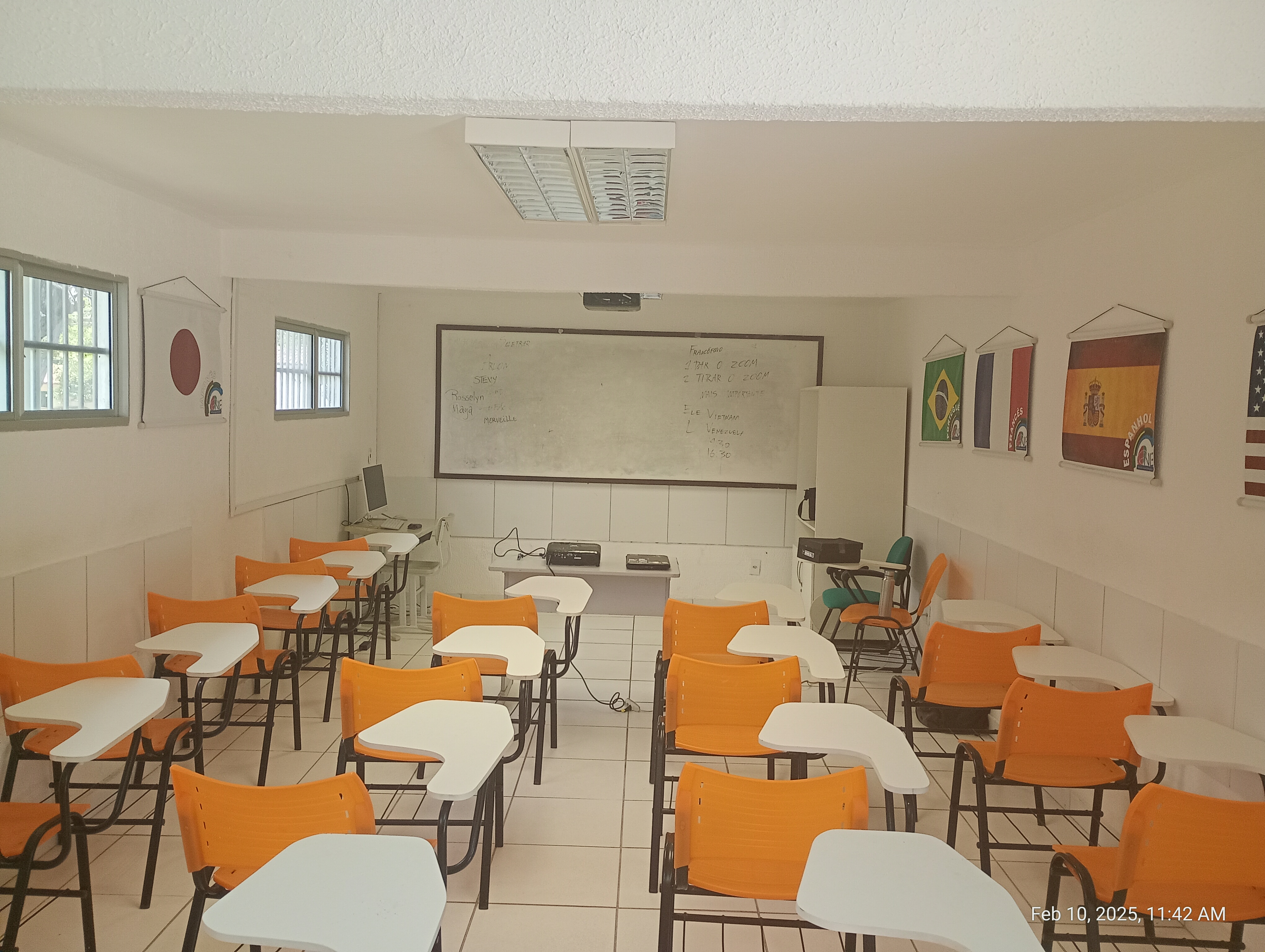 Sala de aula. Várias cadeiras com o assento de cor laranja. Ao fundo , uma lousa para pincel. Ao redor, nas paredes, há bandeiras de vários países.