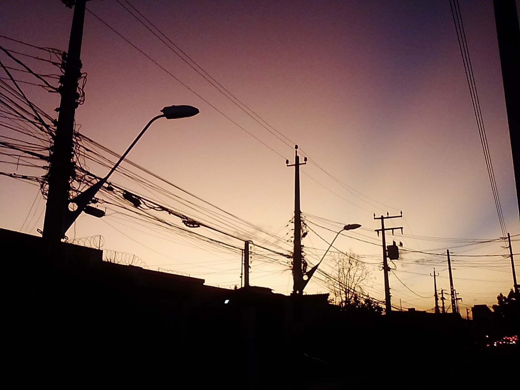 Foto da silhueta de alguns postes de luz contra a luz do final da tarde. No céu, degradês em lilás, amarelo e azul escuro.